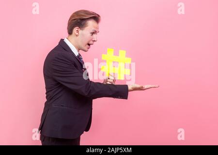 Side view of frustrated shocked man in elegant suit with stylish haircut holding big hashtag symbol and looking in surprise at his palm, opened mouth Stock Photo