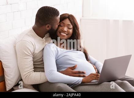 Pregnant afro couple in love chilling out at home, using laptop, laying on bed, free space Stock Photo