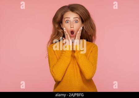 Portrait of extremely excited shocked ginger girl in sweater jumping and holding hands on cheeks and screaming in amazement surprise, her hair flying Stock Photo