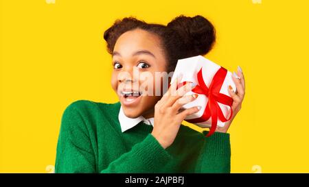 Xmas Present. Curious black teen girl shaking box, checking gift over yellow background, panorama, free space Stock Photo