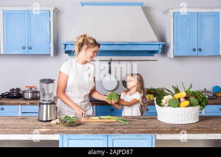 Happy cute mother in apron and little daughter holding broccoli and smiling, cooking salad in kitchen together, preparing vegetarian food, basket of f Stock Photo