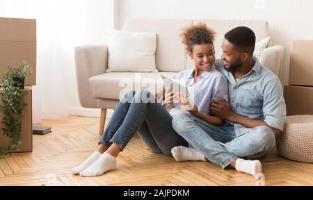 Relocation Concept. Joyful Black Spouses Using Tablet Computer Sitting On Floor Among Moving Boxes In New House. Copy Space Stock Photo