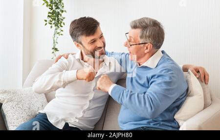 Adult Son And Elderly Father Bumping Fists Sitting On Couch At Home. Relationship With Senior Parents. Selective Focus Stock Photo