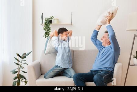 Best Grandpa. Happy Grandfather And Grandson Fighting With Pillows Having Fun Sitting On Sofa At Home. Stock Photo