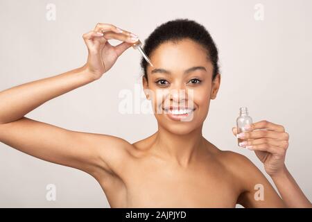 Beauty And Skincare. African American Girl Applying Cosmetic Oil On Face Perfect Skin Standing Over Beige Studio Background Stock Photo