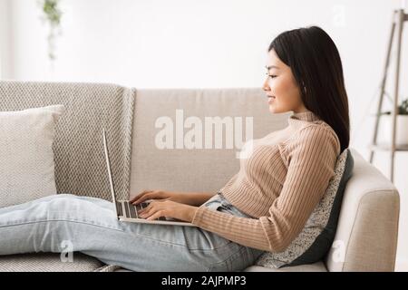 Asian freelancer girl working online on laptop, sitting on comfortable sofa at home, side view Stock Photo