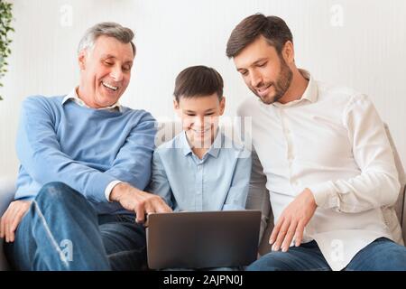 Male Family. Father And Grandfather Helping Boy Do Homework On Laptop Computer Sitting On Sofa At Home Stock Photo