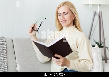 Leisure and literature concept. Middle aged woman reading book and holding glasses, resting at home on sofa Stock Photo