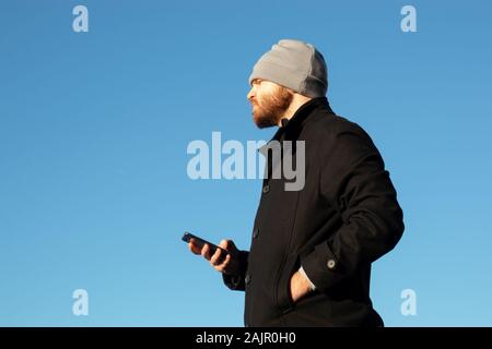 Bearded man with a beanie holding a blue smartphone. Sideview shot with one hand in jacket pocket. Blank blue area of the sky. Looking into the distan Stock Photo
