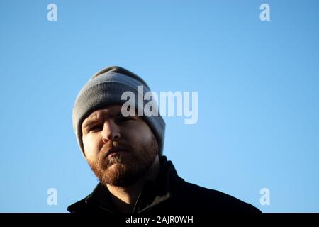 Bearded man with a beanie looking at the camera with a serious look, light blue sky. Serious contemplating thinking mood Stock Photo