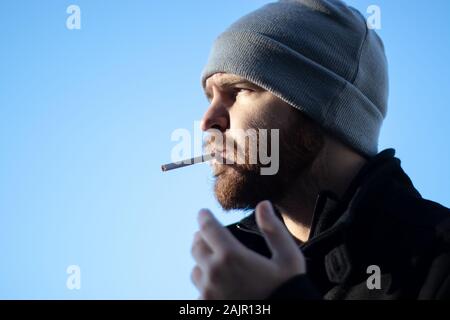 beanie cigarettes Caucasian bearded male with a beanie holding a cigarette, harshing sunlight  with strong shadows. Narrow shot with blue sky background Stock Photo