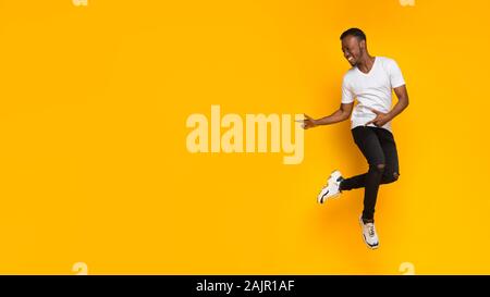 Millennial African American Guy Poniting Fingers Aside Having Fun Jumping And Dancing Over Yellow Studio Background. Copy Space, Panorama Stock Photo