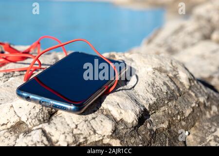 Blue smartphone with red earphone carble on stone wall. Concept of technology. Blue sea in blurred background Stock Photo