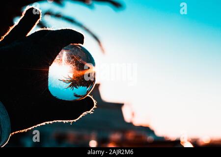 Hand with a glove holding a crystal glass ball, morning harsh sun reflected in the ball. Tones of light blue sky. Stock Photo