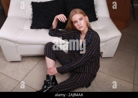 Slim, confident and successful young woman is sitting on the floor dressed in stylish stripped suit and boots (studio shoot of catalogue of fashion Stock Photo