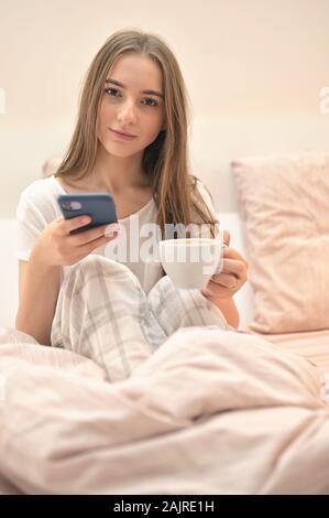 Cheerful Young Woman Sitting In Bed With Smartphone And Cup of Coffee Stock Photo