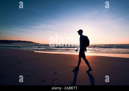 Young man with backpack walking on tropical sand beach. Coast of Sri Lanka at beautiful sunrise.