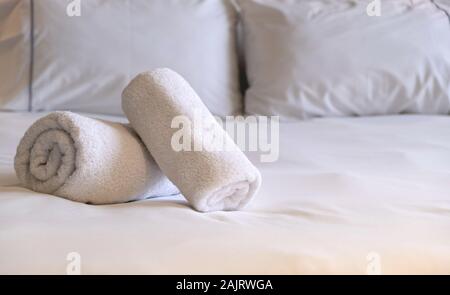 Hotel towels. White fluffy, rolled bath towels, linen sheets and pillows on a bed, bedroom interior detail. Close up view. Stock Photo