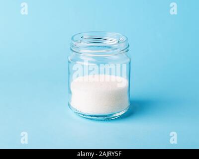 White crystals of sugar in a glass bottle on a pastel blue background. Simple carbohydrate or monosaccharide as food ingredient. Front view. Stock Photo
