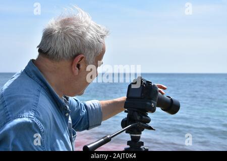 Portrait of a senior man shooting video by the sea with his DSLR camera with telephoto lens on tripod. Out of focus sea at background Stock Photo