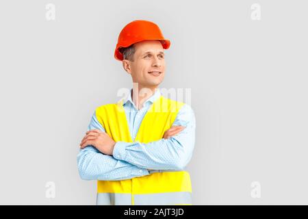Construction. Mature man in hardhat and uniform standing isolated on white crossed arms looking aside dreamful Stock Photo