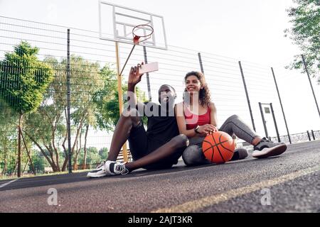 Outdoors Activity. African couple sitting on basketball court taking selfie on smartphone posing to camera cheerful Stock Photo