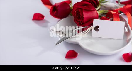 Table setting with a white paper card. White plates, cutlery and decorative textile on white background. Red flowers Stock Photo