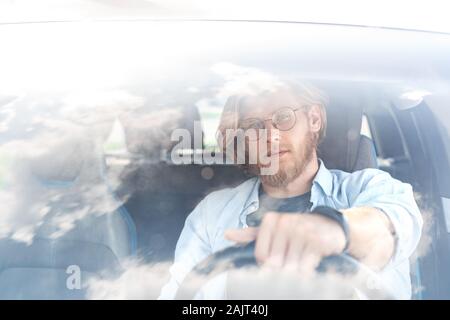 Transportation. Young bearded man in glasses traveling by electric car sitting inside driving looking camera pensive view through windscreen Stock Photo