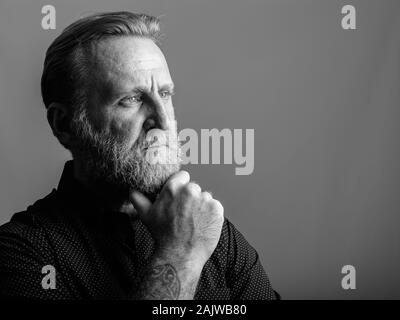 Studio shot of mature bearded man with hand tattoos thinking seriously in gray background Stock Photo