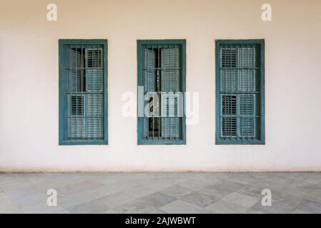 White wall with three grunge windows with wooden green shutters and wrought iron bars and white marble floor Stock Photo