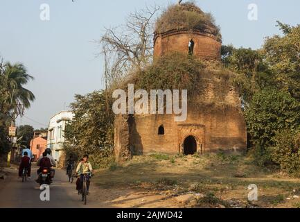 Bishnupur, West Bengal, India - February 6, 2018: An overgrown ruin of an old brick temple built by the Malla dynasty. Kids cycle on the road that goe Stock Photo