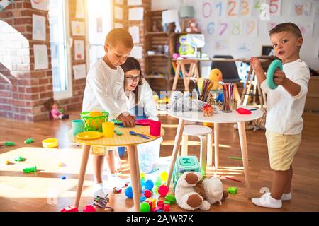 Beautiful teacher and toddlers playing cooking with plastic food around lots of toys at kindergarten Stock Photo