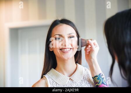 Young black-haired girl looking into camera while getting her face toned with a gentle brush by a pro make up artist preparing her for Valentines Day