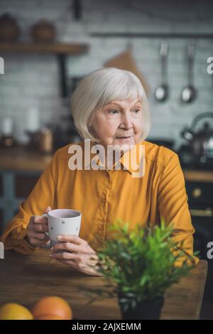 Good-looking elderly lady in yellow sitting in her kitchen Stock Photo