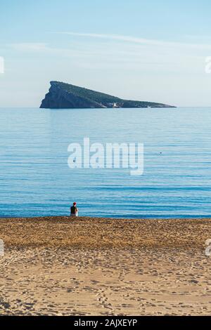 Unrecognizable woman in Santa hat sitting alone in empty beach with Mediterranean sea and Benidorm island background. Spain Stock Photo