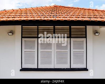 Close up view of the antique wooden window shutters and red tiled roof of a whitewashed aged old British colonial bungalow house in Singapore Stock Photo