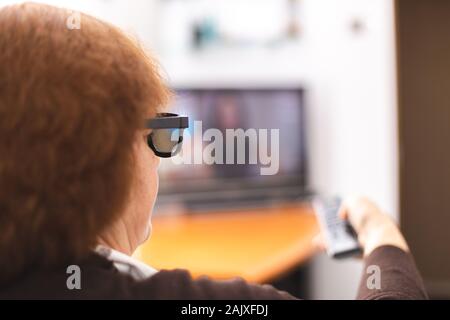 Senior lady with a 3d eyeglasses and tv remote point to a tv screen. Time to watching a tv. Remote control In hand. Focus on eyeglass Stock Photo