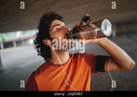 Close-up portrait of a handsome fit young man with earphones in his ears drinking water from a reusable water bottle Stock Photo