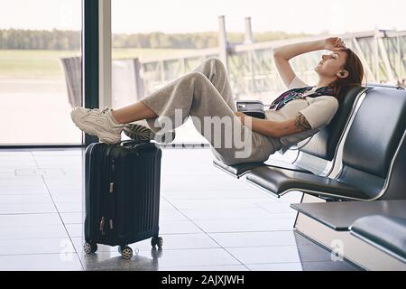 Airport travel lifestyle. Young Gen Z girl traveler sitting in terminal hall with her luggage, trolley bag, passport, smartphone and ticket while Stock Photo