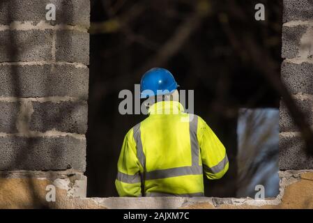 RIGA, LATVIA. 25th March 2019. Selective focus photo. Worker with blue plastic helmet on head and yellow reflecting vest works at building yard. Stock Photo
