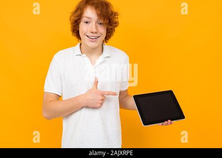 red-haired curly teenager consultant in a store in a white T-shirt shows a tablet display with a mockup on a yellow background Stock Photo