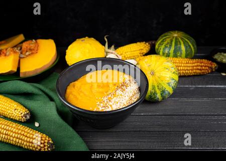 pumpkin cream soup in a black ceramic plate on a dark wooden background using green textile, pieces of pumpkin, seeds, corn. flat lay. copy space Stock Photo