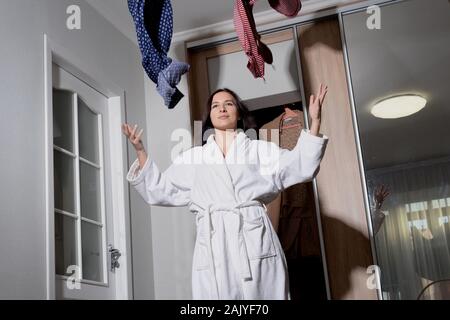 General plan Portrait of a young beautiful girl in a home white coat, sitting on the floor in a room with scattered things, tossing clothes. In the ea Stock Photo