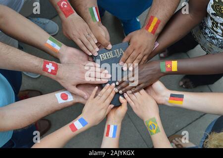 International brothers and sisters in Christ with different flags painted on their arms holding a bible together. Stock Photo