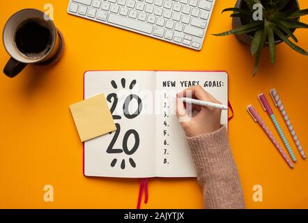 Stock photo of a young woman hand writing in a 2020 new year notebook with list of resolutions and objects on yellow background Stock Photo