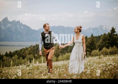 happy bride and groom walk through field in front of mountains Wyoming Stock Photo