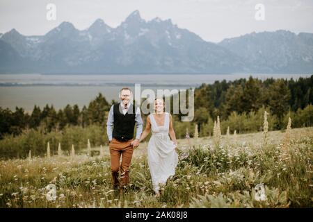 happy smiling newlyweds walk through field of wildflowers in Tetons Stock Photo