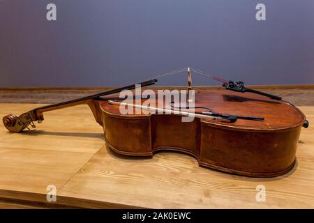 An old broken double bass lying on a wooden table on the background of a wall. Stock Photo