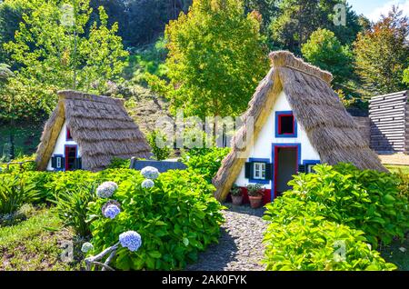 Santana, Madeira, Portugal - Sep 24, 2019: Traditional Madeiran houses with straw roofs in the Madeira Theme Park. Typical old buildings surrounded by green trees and Hydrangea flowers. Stock Photo