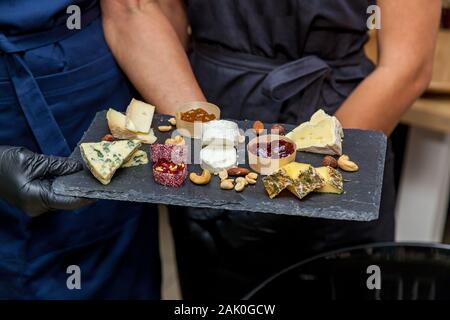 A cheese plate with an assortment of various types of cheese on a dark board on the background of tasters. Stock Photo